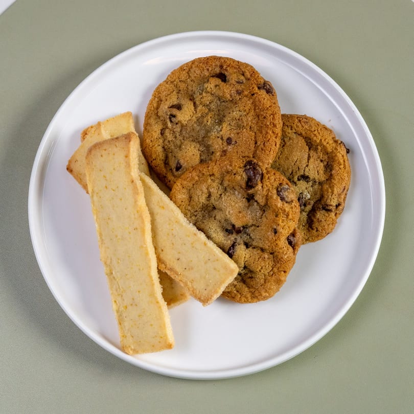 A white plate holds three chocolate chip cookies and four shortbread fingers on a green surface.