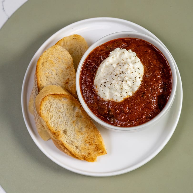 A white bowl of red chili with a dollop of white sour cream and black pepper, served with toasted bread slices.