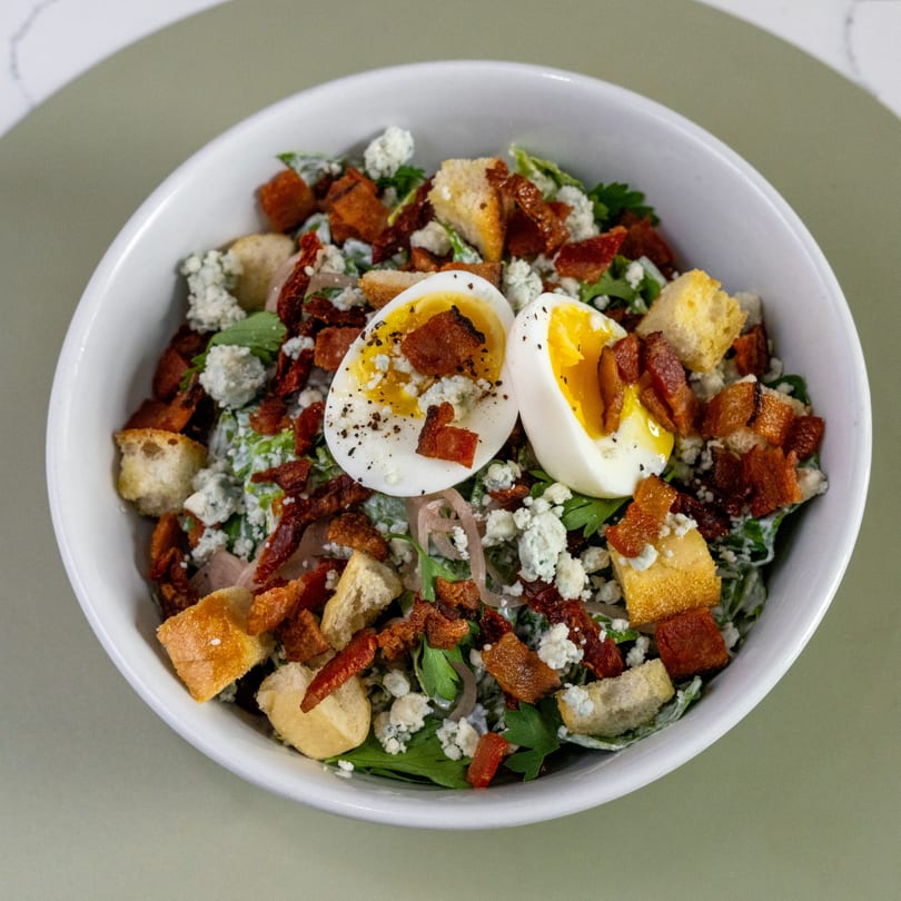 Overhead view of a salad with greens, bacon, croutons, blue cheese, and two soft-boiled egg halves.